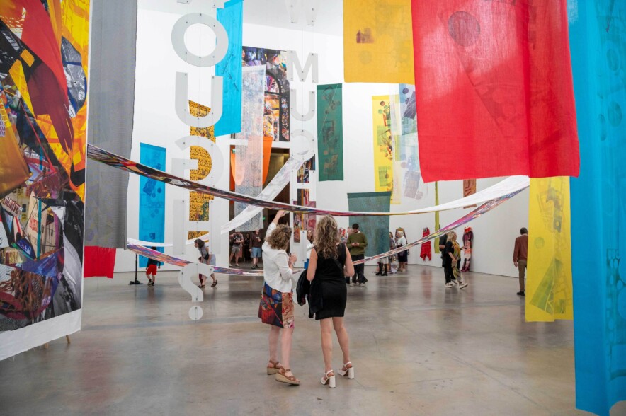 Gallery visitors in a large, bright space looking at art hanging from the ceiling.